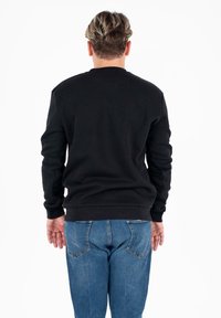 Uomo con capelli corti e mossi che indossa una maglietta nera a maniche lunghe e jeans blu, in piedi di spalle su uno sfondo bianco.