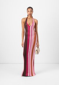 Strikket maxikjole med vertikale striper i nyanser av rosa, brunt og oransje, med dyp V-hals og sideslitt. Modellen holder en liten beige veske.