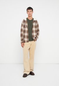 Veste en flanelle à carreaux marron avec une capuche doublée de blanc, portée sur un T-shirt henley vert. Pantalon large beige clair et chaussures marron foncé.