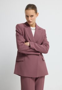 Op maat gemaakte blazer van zacht mauve stof, met scherpe revers, twee voorkezen en een ontspannen pasvorm. Gedragen over een witte top met versieringen.