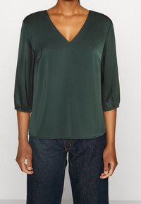 Dunkelgrüne Satin-Bluse mit V-Ausschnitt, dreiviertel Ärmeln und lässiger Passform. Glatte Textur mit minimalen Nähdetails.