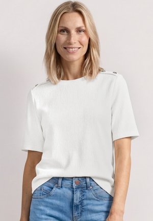 Femme souriante aux cheveux blonds mi-longs, portant une chemise blanche texturée à manches courtes avec des boutons aux épaules et un jean bleu.