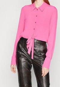 Blusa rosa con bottoni, colletto, maniche lunghe e dettaglio anteriore annodato. Abbinata a pantaloni in pelle nera lucida, che mostrano una silhouette elegante.