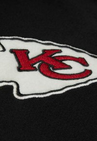 Rote KC-Buchstaben auf einem weißen, pfeilförmigen Patch auf schwarzem Stoffhintergrund gestickt.