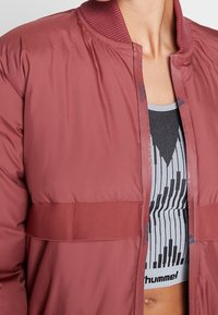 Chaqueta acolchada burdeos con cuello acanalado y cremallera frontal. Presenta una amplia banda horizontal en el pecho. Superpuesta a una camiseta con estampado negro y gris.