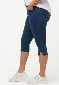 Pantalones capri de mezclilla en azul oscuro con una textura suave, bolsillos laterales y dobladillos divididos, combinados con unas zapatillas deportivas de color claro.