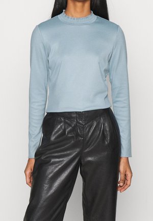 Helles blaues Langarmshirt mit gerafftem Ausschnitt, kombiniert mit hochgeschnittenen schwarzen Lederhosen, die eine glatte Textur und einen taillierten Schnitt aufweisen.