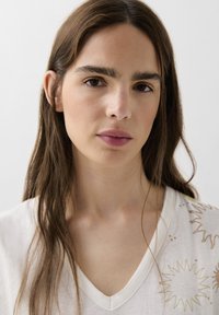 Light-colored, cotton V-neck top with cream fabric, featuring gold sunburst embroidery on one side. Smooth texture and relaxed fit.