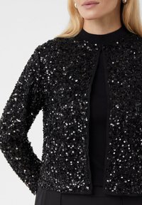 Kobieta w czarnej kurtce z cekinami, zapinanej z przodu, nosi czarną bluzkę z wysokim kołnierzem, o prążkowanej fakturze. Jej długie blond włosy są widoczne.