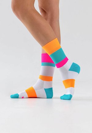 Beine an den Knöcheln gekreuzt, weiße Socken mit breiten horizontalen Streifen in Orange, Türkis und knalligem Pink auf hellem Hintergrund.