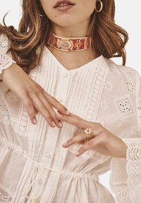 Blusa blanca bordada con cierre de botones, que presenta un delicado borde de encaje y una cintura ajustable con cordón. La modelo lleva un gargantilla estampada y un anillo.