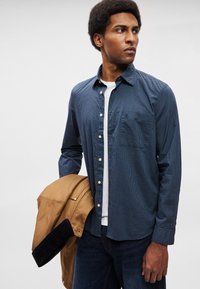 Μπλε καρό πουκάμισο με κουμπιά και τσέπη στο στήθος, φορεμένο πάνω από ένα λευκό μπλουζάκι. Κρατώντας μια καφέ και μαύρη ζακέτα. Σκούρα τζιν.