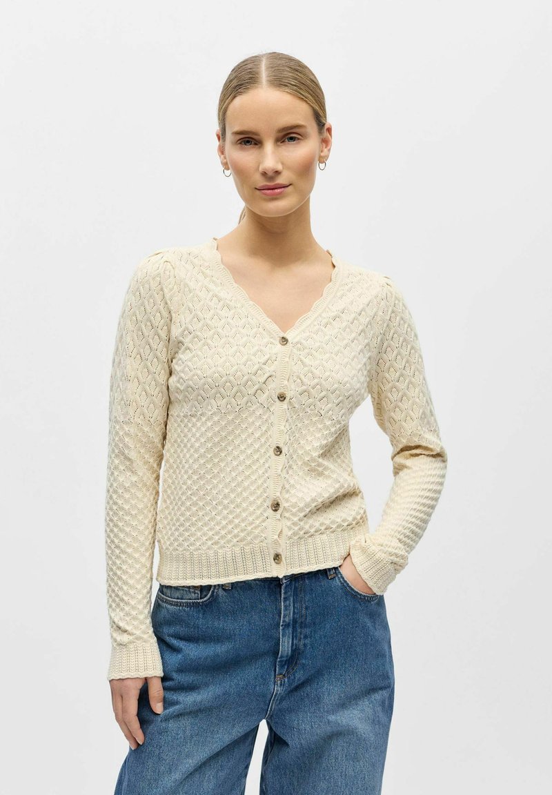 Femme portant un cardigan crème texturé boutonné et un jean bleu, debout avec une main dans la poche devant un fond uni.