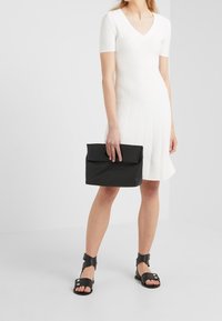 Robe blanche côtelée à manches courtes avec un col en V. Pochette noire tenue dans une main, associée à des sandales noires à brides autour de la cheville.