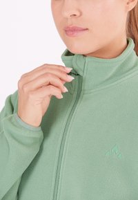 Grüne Fleecejacke mit hohem Kragen und Reißverschluss, mit einem Logo auf der Brust. Weicher Stoff, figurbetonte Passform und elastische Bündchen.