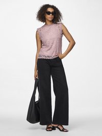 Kvinne med krøllete hår som har på seg svarte solbriller, ermeløs rosa bluse i blonder, vide svarte bukser, svarte hæler, og holder en stor svart veske.