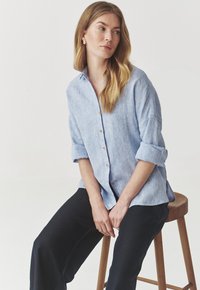 Camisa de lino azul claro con botones en el frente, mangas enrolladas y un corte relajado, combinada con pantalones negros.