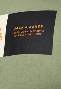 T-shirt in cotone verde con una bold grafica rettangolare nera contenente testo bianco e arancione, inclusi dettagli sul marchio e sulla località.