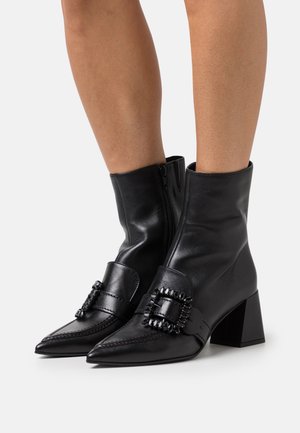 Botas de tobillo de cuero negro con punta afilada, cierre de cremallera y detalle de hebilla decorativa. Tacones cuadrados gruesos, textura suave.