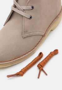 Beige suede ankle boot with light brown stitching and two brown laces beside it. Boot features black eyelets and a simple design.