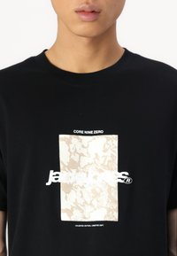 Sort bomulds T-shirt med et stort cremefarvet grafisk mønster og hvid tekst med påskriften "jack & jones" og "CORE NINE ZERO."
