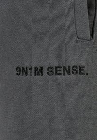 Grå bomullstoff med brodert tekst "9N1M SENSE." med en jevn tekstur og et subtilt strikkemønster. Synlig kantsting.