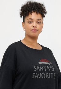 Melns džemperis ar uzrakstu "SANTA'S FAVORITE" ietērptu rhinestone, papildināts ar sarkanu un baltu Ziemassvētku vecīša cepuri. Mīksta tekstūra, brīvs griezums, apaļa dekoltē.