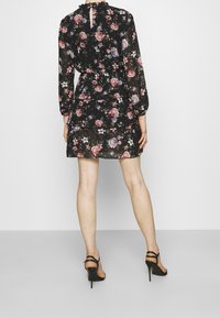 Robe noire à motifs floraux avec manches longues, design en couches, ornée de roses roses et violettes. Portée avec des sandales noires à talons hauts.