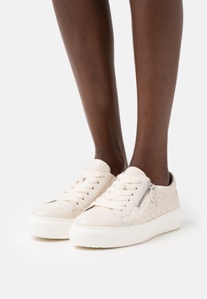 Dark-skinned legs wearing cream-colored sneakers with lace-up fronts, quilted sides, and silver zipper detail on a white background.