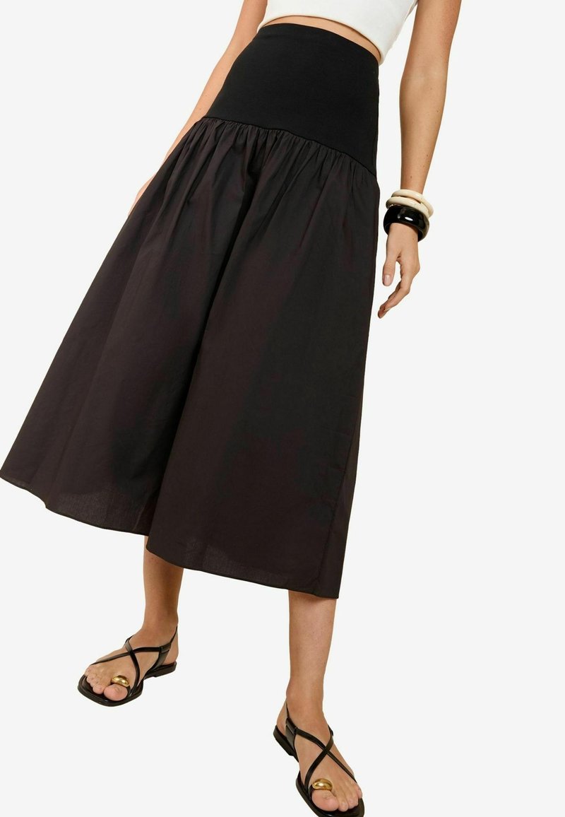 Mustaherra, polvimittainen hame, jossa on leveä, joustava musta vyötärö ja rypytetty kangas. Yhdistetty mustiin sandaaleihin, joissa on kultainen yksityiskohta.