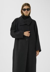 Överdimensionerad svart ullrock med bred krage, enknappsstängning och lösa ärmar. Bärs med en svart polotröja och solglasögon.
