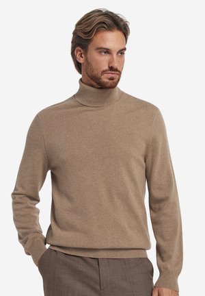 Mann mit hellbraunen Haaren und Bart, der einen beigen Rollkragenpullover und braune Hose trägt und mit einer Hand in der Tasche zur Seite schaut.
