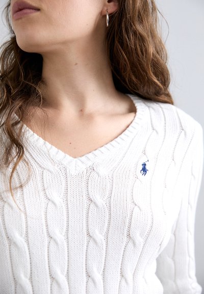 Pull en maille blanche avec un col en V et un motif en chevrons. Dispose d'un petit logo bleu sur la poitrine gauche et de poignets côtelés. Texture douce.