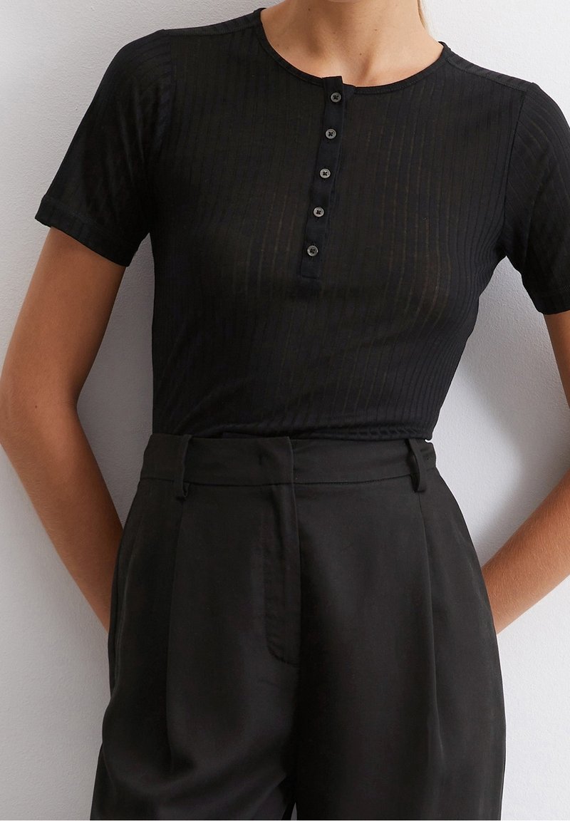 Femme portant un t-shirt Henley noir côtelé à manches courtes et ajusté, avec cinq boutons, rentré dans un pantalon noir taille haute.