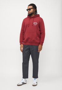 Hombre con gafas de sol, sudadera roja con bolsillo frontal, pantalones gris oscuro y zapatillas Adidas blancas, de pie contra un fondo sencillo.