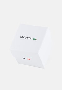 Caja cuadrada blanca con el logotipo de Lacoste en negro y verde, con dos acentos de color: uno negro y otro rojo en el lado frontal.
