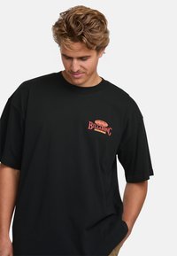 Svart kortärmad t-shirt i bomull. Har en röd Billabong-logotyp på vänster bröst och en lös passform. Slät textur, enkel design.