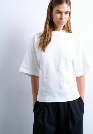 Jeune femme aux longs cheveux châtain clair, portant un haut blanc ample à manches courtes et un pantalon noir, debout devant un fond clair uni.