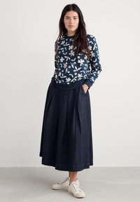 Tengerészi pulóver fehér virágmintával és bordás mandzsettával, hosszú sötét farmer szoknyával párosítva, amely elöl redőzött és oldzsebes. Fehér sportcipő.