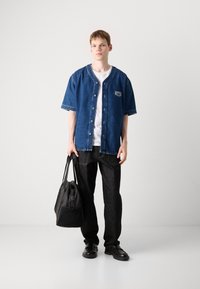 Denimskjorte i mørkeblå med korte ermer og trykknapper, over en hvit t-skjorte. Svarte jeans og en svart skulderveske fullfører antrekket.