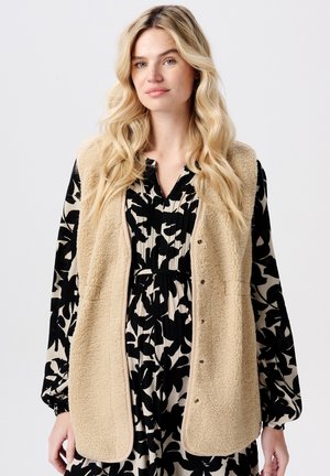 Vrouw met lang blond haar, gekleed in een beige fleece vest over een zwart en crèmekleurig bloemenpatroon jurk, staand tegen een effen achtergrond.