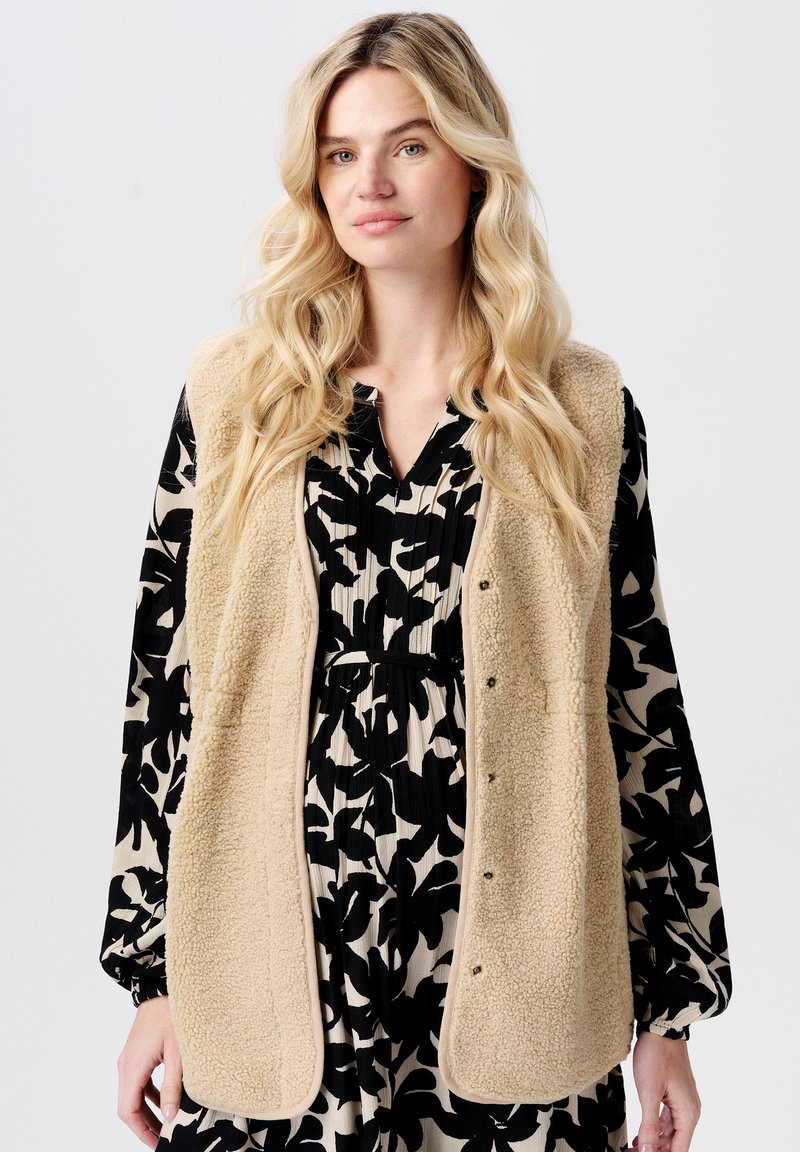 Frau mit langen blonden Haaren, die eine beigem Fleece-Weste über einem schwarz-cremefarbenen, floral gemusterten Kleid trägt, steht vor einem schlichten Hintergrund.