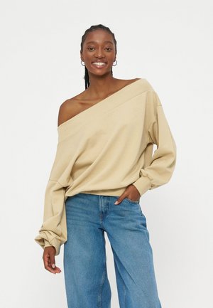 Mosolygó nő fonott hajjal bézs váll nélküli pulóvert és kék farmert visel, egyik kezét a zsebében tartva egy fehér háttér előtt áll.