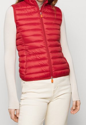 Woman wearing a red quilted sleeveless vest over a beige long-sleeve top and off-white pants against a plain background.