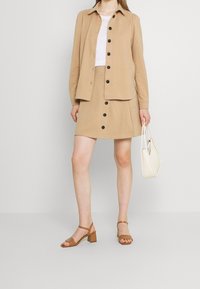 Giacca beige con bottoni neri, gonna abbinata, sandali con tacco marroni e una borsa bianca. Il tessuto sembra morbido e ha una texture liscia.