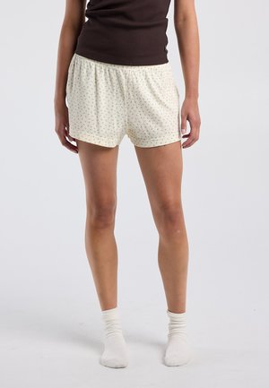 Femme portant un short couleur crème avec un petit motif floral, un haut sans manches marron foncé et des chaussettes blanches aux chevilles, debout sur un fond uni.