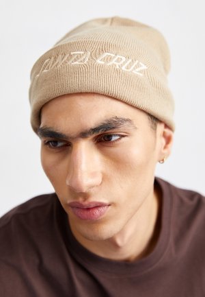 Jeune homme portant un bonnet en tricot beige avec l'inscription "Santa Cruz", une chemise foncée et une petite boucle d'oreille en anneau, regardant légèrement vers le bas.