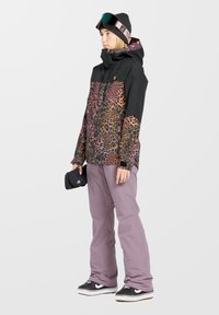 Färgglad leopardmönstrad snöjacka med svart överdel och rosa detaljer, kombinerad med ljuslila snöbyxor och svarta sneakers.