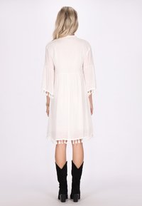 Weißes Kleid mit Dreiviertel-Bellärmeln und Quastenakzenten am Saum. Leichter Stoff und eine lockere, fließende Silhouette.