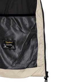 Giacca isolante tan e nera con interno in rete traspirante, etichetta nera con testo dorato che evidenzia caratteristiche come la protezione dall'acqua.